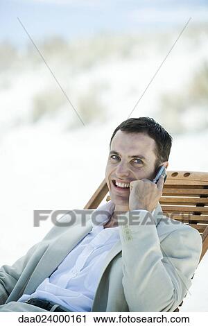Stock Image - Businessman sitting in deck chair at beach, using cell phone. Fotosearch
