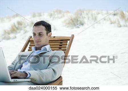Businessman sitting in deck chair at beach, using laptop View Large Photo Image Stock Photography - Businessman sitting in deck chair at beach, using laptop. Fotosearch