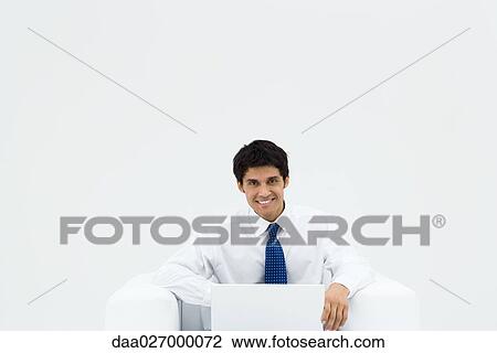 Businessman sitting on armchair using laptop, smiling at camera View Large Photo Image Stock Image - Businessman sitting on armchair using laptop, smiling at camera. Fotosearch