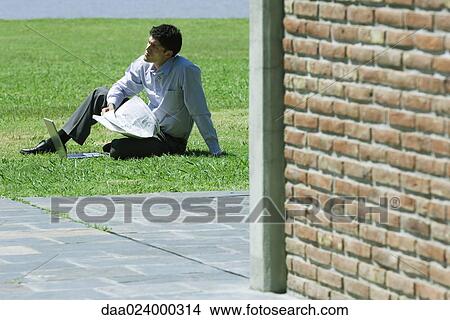 Businessman sitting on grass with laptop and newspaper, corner of brick building in foreground View Large Photo Image Picture - Businessman sitting on grass with laptop and newspaper, corner of brick building in foreground. Fotosearch