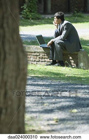 Stock Image - Businessman sitting on low wall in park, using laptop. Fotosearch