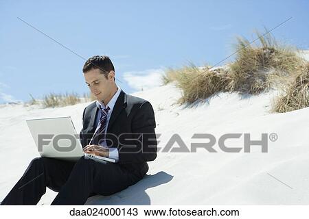 Businessman sitting on sand dune, using laptop View Large Photo Image Stock Image - Businessman sitting on sand dune, using laptop. Fotosearch
