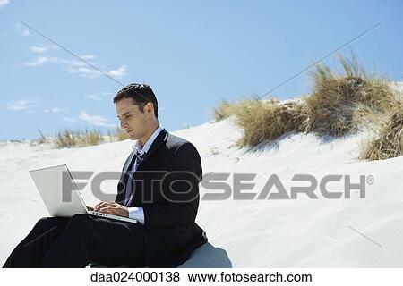Businessman sitting on sand dune, using laptop View Large Photo Image Stock Photo - Businessman sitting on sand dune, using laptop. Fotosearch