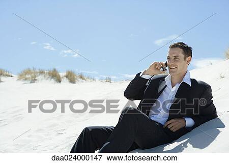 Stock Photo - Businessman sitting on sand dune, using cell phone. Fotosearch