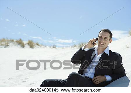 Stock Photography - Businessman sitting on sand dune, using cell phone. Fotosearch