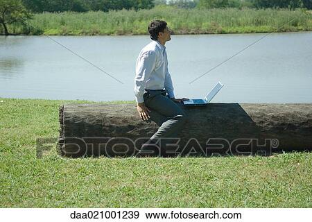 Businessman sitting on tree trunk with laptop, looking away View Large Photo Image Stock Photo - Businessman sitting on tree trunk with laptop, looking away. Fotosearch