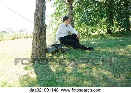 Businessman sitting under tree on bench, with laptop View Large Photo Image Stock Photo - Businessman sitting under tree on bench, with laptop. Fotosearch