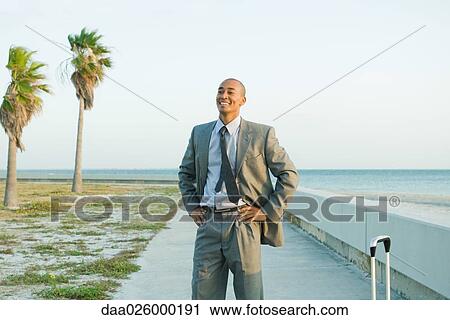Stock Image - Businessman standing at the beach with hands on hips, smiling, looking away. Fotosearch