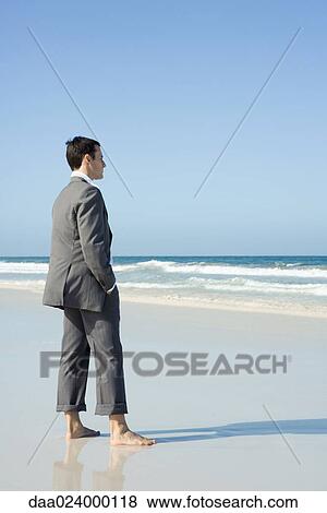Stock Photo - Businessman standing barefoot on beach with hands in pockets, side view. Fotosearch