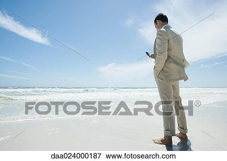 Businessman standing barefoot on beach, holding cell phone View Large Photo Image Stock Photo - Businessman standing barefoot on beach, holding cell phone. Fotosearch