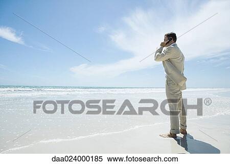 Stock Photography - Businessman standing barefoot on beach, using cell phone. Fotosearch