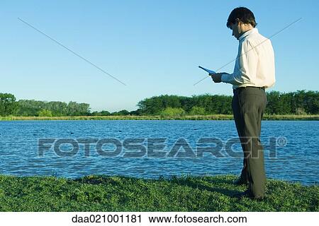 Stock Image - Businessman standing by edge of water, reading documents. Fotosearch