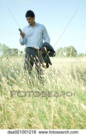 Businessman standing in field, looking at cell phone View Large Photo Image Stock Photo - Businessman standing in field, looking at cell phone. Fotosearch