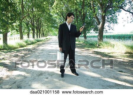 Businessman standing in rural road, holding briefcase, looking at cell phone View Large Photo Image Stock Photo - Businessman standing in rural road, holding briefcase, looking at cell phone. Fotosearch