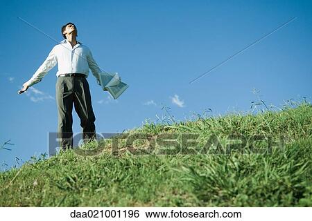 Stock Photograph - Businessman standing on grassy hill, arms out, head back and eyes closed, low angle view. Fotosearch