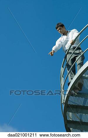 Stock Image - Businessman standing on metal balcony, pointing down, low angle view, blue sky in background. Fotosearch