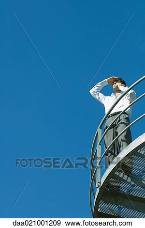 Businessman standing on metal balcony, looking away, low angle view, blue sky in background View Large Photo Image Stock Photo - Businessman standing on metal balcony, looking away, low angle view, blue sky in background. Fotosearch