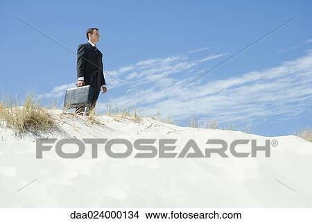 Businessman standing on sand dune, holding briefcase, low angle view View Large Photo Image Picture - Businessman standing on sand dune, holding briefcase, low angle view. Fotosearch