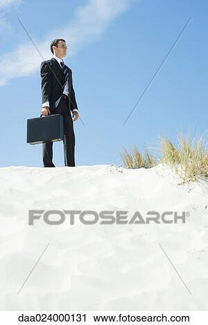 Businessman standing on sand dune, holding briefcase, low angle view View Large Photo Image Stock Image - Businessman standing on sand dune, holding briefcase, low angle view. Fotosearch