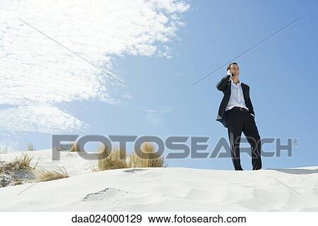 Businessman standing on sand dune, using cell phone, low angle view View Large Photo Image Stock Photo - Businessman standing on sand dune, using cell phone, low angle view. Fotosearch