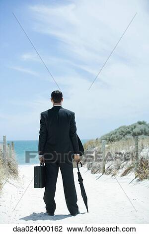 Businessman standing on sandy path leading to ocean, holding briefcase and umbrella, rear view View Large Photo Image Stock Image - Businessman standing on sandy path leading to ocean, holding briefcase and umbrella, rear view. Fotosearch