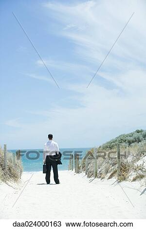 Businessman standing on sandy path leading to ocean, rear view View Large Photo Image Stock Image - Businessman standing on sandy path leading to ocean, rear view. Fotosearch