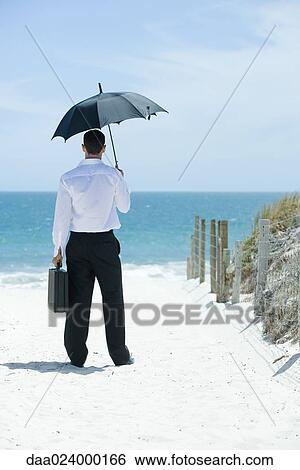 Businessman standing under umbrella on sandy path leading to ocean, rear view View Large Photo Image Stock Photograph - Businessman standing under umbrella on sandy path leading to ocean, rear view. Fotosearch