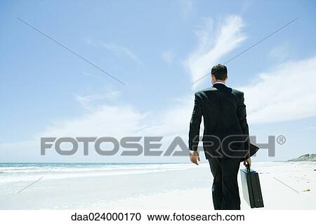 Stock Image - Businessman walking across beach, carrying briefcase, rear view. Fotosearch