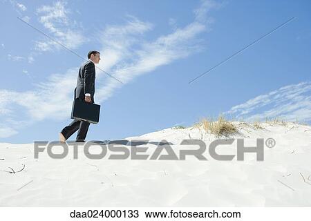 Stock Image - Businessman walking across sand dune, low angle view. Fotosearch