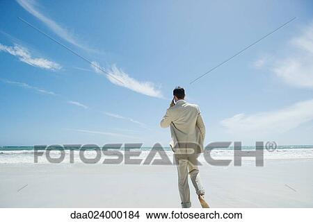 Picture - Businessman walking barefoot on beach, using cell phone. Fotosearch
