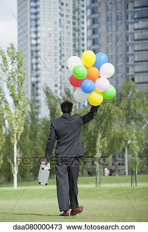 Stock Image - Businessman walking with bunch of balloons, rear view. Fotosearch