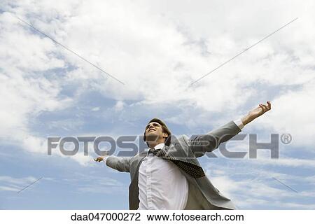 Stock Image - Businessman with arms outstretched against cloudy sky. Fotosearch