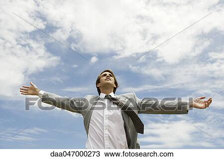 Stock Image - Businessman with arms outstretched against cloudy sky. Fotosearch