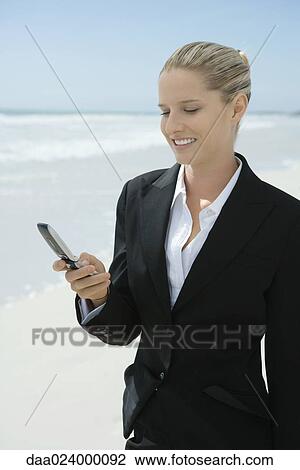 Stock Image - Businesswoman holding cell phone, standing on beach, smiling. Fotosearch