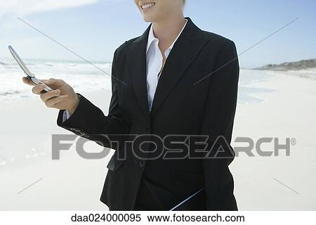 Stock Photography - Businesswoman holding cell phone, standing on beach, smiling, cropped. Fotosearch