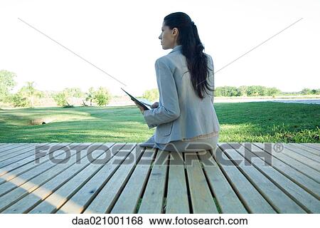 Stock Photo - Businesswoman sitting on edge of deck, rear view. Fotosearch