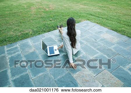 Stock Photograph - Businesswoman sitting on patio with laptop, looking away, high angle, rear view. Fotosearch