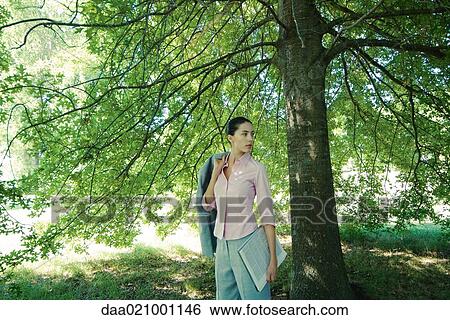 Stock Photograph - Businesswoman standing under tree, holding jacket over shoulder, looking away. Fotosearch