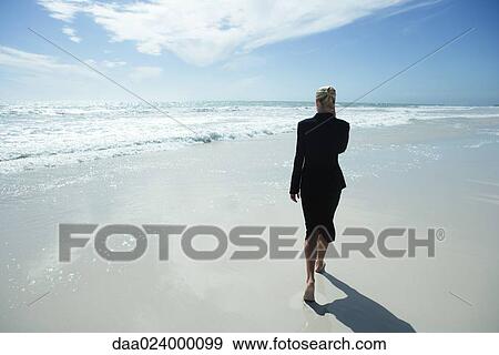 Stock Photo - Businesswoman using cell phone, walking barefoot across beach, full length, rear view. Fotosearch