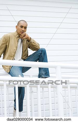 Picture - Casually dressed man sitting on railing, hand under chin, looking at camera. Fotosearch