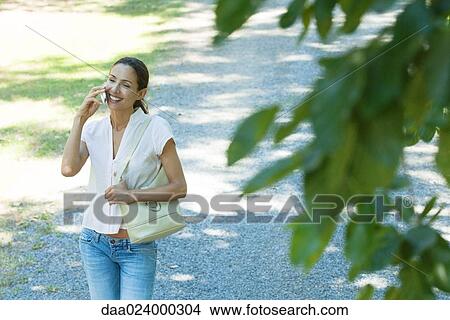 Casually dressed woman using cell phone, smiling as she walks through park, purse on shoulder View Large Photo Image Picture - Casually dressed woman using cell phone, smiling as she walks through park, purse on shoulder. Fotosearch