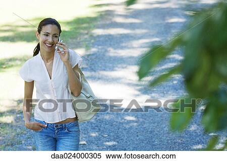 Casually dressed woman using cell phone, smiling as she walks through park, purse on shoulder View Large Photo Image Stock Photography - Casually dressed woman using cell phone, smiling as she walks through park, purse on shoulder. Fotosearch
