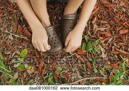 Stock Image - Child sitting on the ground, touching plastic clogs, cropped. Fotosearch
