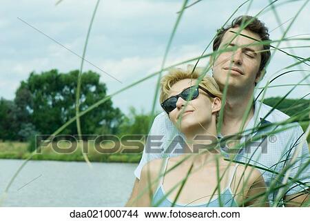 Couple by lake, woman wearing sunglasses, man closing eyes, portrait View Large Photo Image Picture - Couple by lake, woman wearing sunglasses, man closing eyes, portrait. Fotosearch