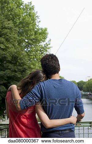 Stock Photo - Couple embracing on bridge, looking at view. Fotosearch