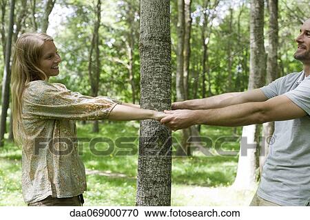 Stock Image - Couple holding hands around tree trunk. Fotosearch