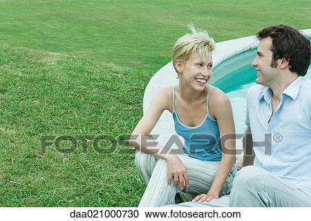 Stock Image - Couple sitting by edge of pool, looking at each other, smiling. Fotosearch