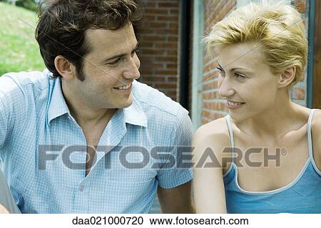 Stock Image - Couple sitting in backyard smiling at each other, brick house in background, close-up. Fotosearch