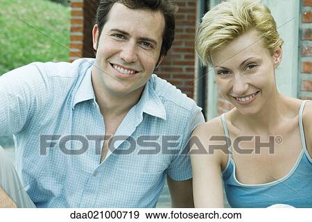 Stock Photo - Couple sitting in backyard smiling at camera, brick house in background, close-up. Fotosearch