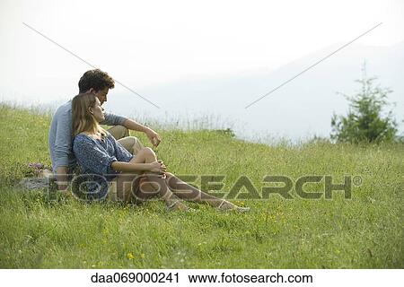 Stock Image - Couple sitting on meadow looking at view. Fotosearch